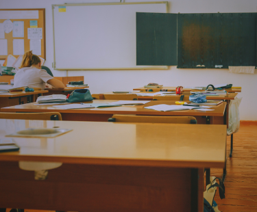 A nearly empty classroom with scattered papers and supplies on desks. A single teacher sits at the back, head down and working, suggesting fatigue or deep focus.